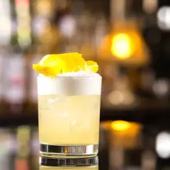 Whisky Sour z pianką i skórką cytryny w szklance na tle rozmytego baru.