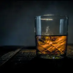 Szklanka bursztynowego trunku z lodem na drewnianym blacie. Z czego robi się whisky? Z ziaren zbóż, wody i drożdży, a potem leżakuje w beczkach.