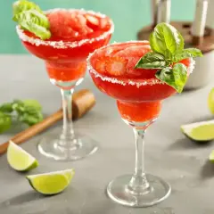Dwa mrożone daiquiri truskawkowe w kieliszkach z cukrowym rantem, ozdobione bazylią i truskawkami.