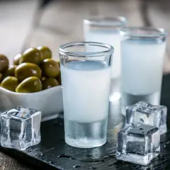 Trzy kieliszki uzo z lodem i miseczka oliwek na drewnianym stole.