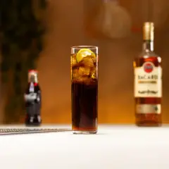 Szklanka z drinkiem z rumem Bacardi, lodem i limonką, obok butelka Coca-Coli i rumu.