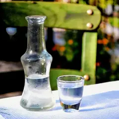 Szklana butelka i kieliszek z przezroczystym alkoholem na białym obrusie, w tle zielone krzesło i rośliny.