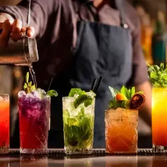 Mistrz miksologii przygotowuje kolorowe drinki. Na blacie baru stoją drinki z owocami i miętą, a barman nalewa płyn z shakera.