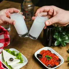 Dwie szklanki z białym alkoholem, raki, unoszą się do toastu. Na stole ser, sałatka i butelka Yeni Raki.