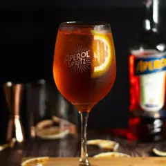 Aperol Spritz w kieliszku z plasterkiem pomarańczy, obok butelka Aperol i inne dodatki.
