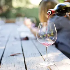 Czerwone wino Carlo Rossi nalewane do kieliszka na drewnianym stole, w tle rozmyte postacie i zieleń.
