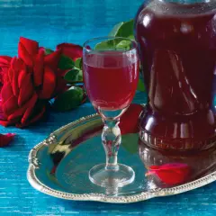 Aromatic nalewka z płatków róży w kieliszku i karafce, obok czerwonej róży i płatków na srebrnej tacy.