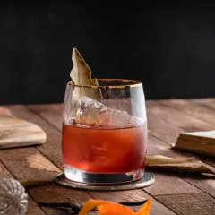 Czerwony koktajl z lodem i liściem laurowym w szkle, obok karafki i starej książki. To może być drink z wermutem.