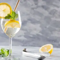Szklanka z drinkiem Hugo, plasterkami cytryny, miętą i słomką. Obok leży połówka cytryny i szczypce.