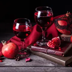 Dwa kieliszki czerwonego wina, granaty i maska tworzą tajemniczą kompozycję. Wino z granatu kusi głębokim kolorem.