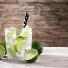 Szklanka z drinkiem, lodem i plasterkami limonki, obok kilka całych limonek.