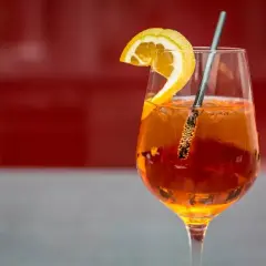 Szklanka Aperol Spritz z plasterkiem pomarańczy i metalową słomką na czerwonym tle.