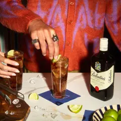 Dwie osoby przygotowują drinki z whisky i colą, z butelką Ballantine's, lodem i limonkami.