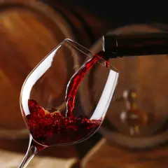 Czerwone wino, bogate w taniny, leje się z butelki do kieliszka, na tle drewnianych beczek.