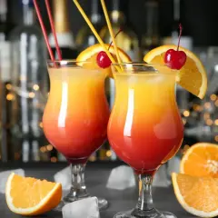Dwa drinki z pomarańczą i wiśnią, na tle butelek i światełek.