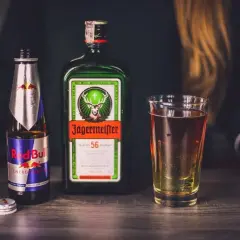 Butelka Jägermeister, puszka Red Bull i szklanka z drinkiem na drewnianym blacie.