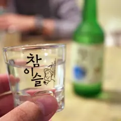 Szklanka z soju z koreańskim napisem i rysunkiem, w tle zielona butelka.