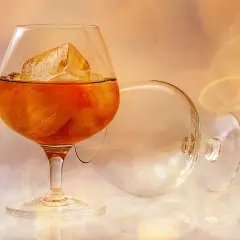 Szklanka z bursztynową brandy i lodem, obok pusta, przewrócona kieliszka.
