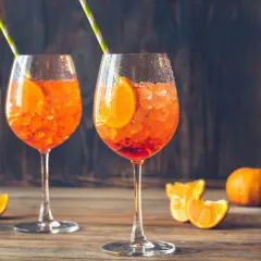 Dwa kieliszki Aperol Spritz z lodem i plasterkami pomarańczy na drewnianym stole.