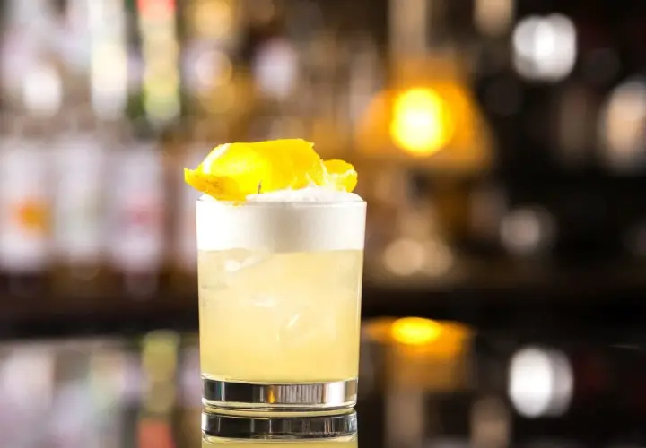 Whisky Sour z pianką i skórką cytryny w szklance na tle rozmytego baru.