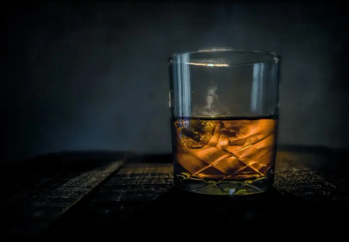 Szklanka bursztynowego trunku z lodem na drewnianym blacie. Z czego robi się whisky? Z ziaren zbóż, wody i drożdży, a potem leżakuje w beczkach.