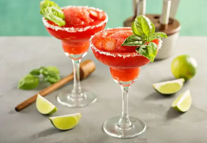 Dwa mrożone daiquiri truskawkowe w kieliszkach z cukrowym rantem, ozdobione bazylią i truskawkami.