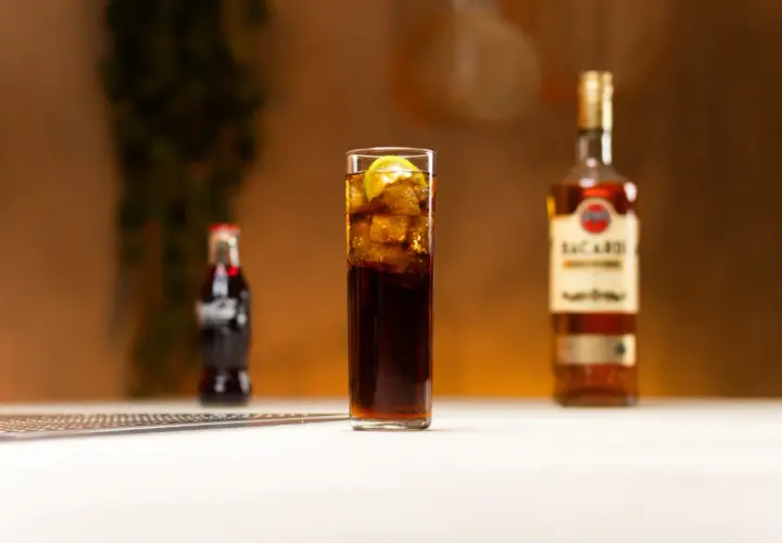 Szklanka z drinkiem z rumem Bacardi, lodem i limonką, obok butelka Coca-Coli i rumu.
