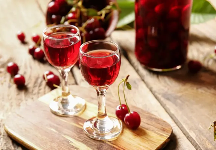 Dwa kieliszki z rubinowym winem z czereśni, obok świeże owoce i słoik pełen wiśni.