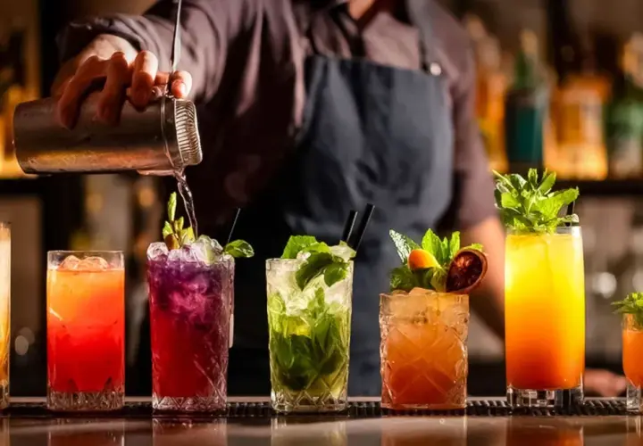 Mistrz miksologii przygotowuje kolorowe drinki. Na blacie baru stoją drinki z owocami i miętą, a barman nalewa płyn z shakera.