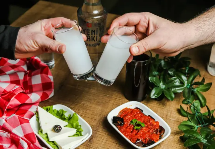 Dwie szklanki z białym alkoholem, raki, unoszą się do toastu. Na stole ser, sałatka i butelka Yeni Raki.
