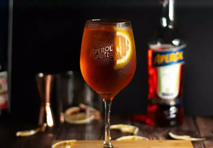 Aperol Spritz w kieliszku z plasterkiem pomarańczy, obok butelka Aperol i inne dodatki.