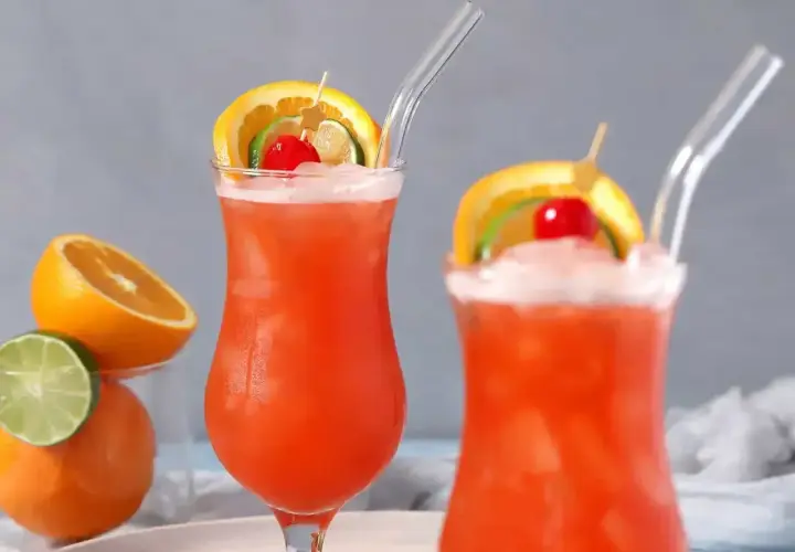 Dwa drinki z rumem, ozdobione plasterkami pomarańczy, limonki i wiśnią, z owocami w tle.