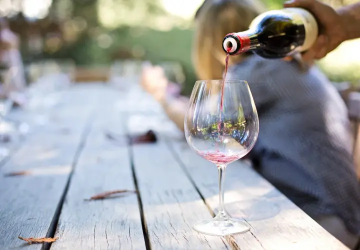Czerwone wino Carlo Rossi nalewane do kieliszka na drewnianym stole, w tle rozmyte postacie i zieleń.