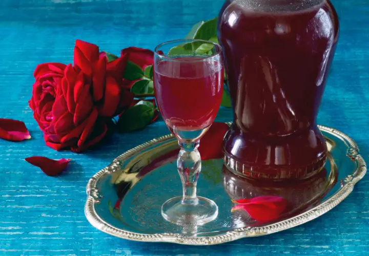Aromatic nalewka z płatków róży w kieliszku i karafce, obok czerwonej róży i płatków na srebrnej tacy.