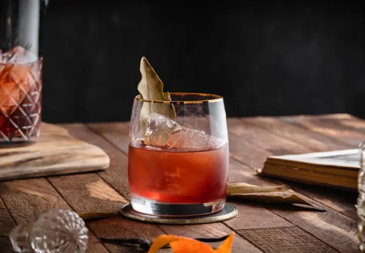 Czerwony koktajl z lodem i liściem laurowym w szkle, obok karafki i starej książki. To może być drink z wermutem.
