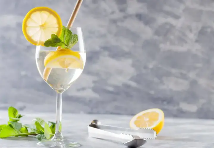 Szklanka z drinkiem Hugo, plasterkami cytryny, miętą i słomką. Obok leży połówka cytryny i szczypce.