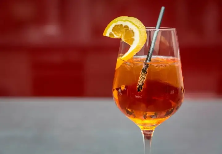 Szklanka Aperol Spritz z plasterkiem pomarańczy i metalową słomką na czerwonym tle.