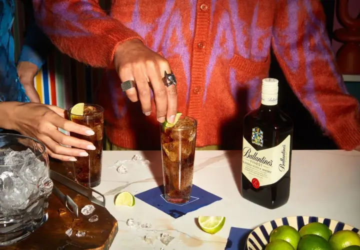 Dwie osoby przygotowują drinki z whisky i colą, z butelką Ballantine's, lodem i limonkami.