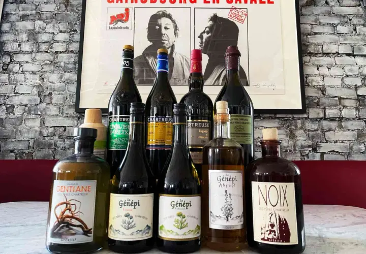 Butelki z zielonymi likierami Chartreuse i innymi francuskimi alkoholami na tle plakatu.