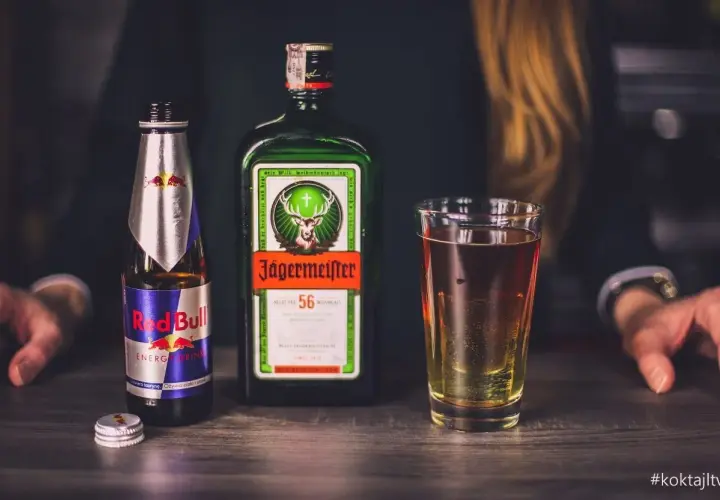 Butelka Jägermeister, puszka Red Bull i szklanka z drinkiem na drewnianym blacie.