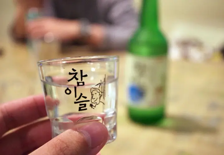 Szklanka z soju z koreańskim napisem i rysunkiem, w tle zielona butelka.