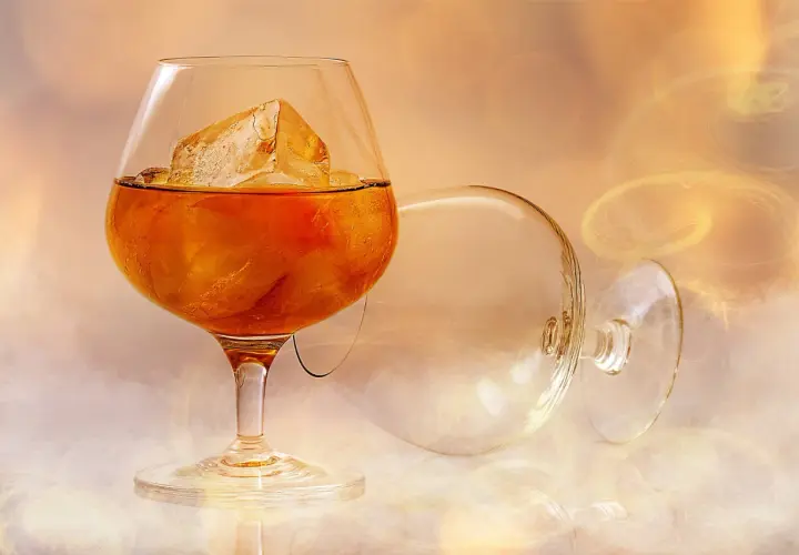 Szklanka z bursztynową brandy i lodem, obok pusta, przewrócona kieliszka.