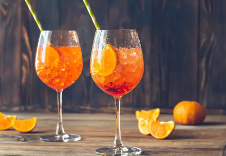 Dwa kieliszki Aperol Spritz z lodem i plasterkami pomarańczy na drewnianym stole.