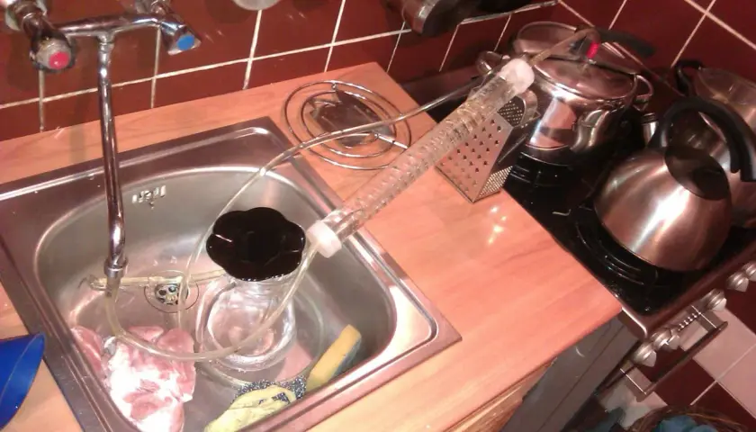 Zlew kuchenny z kranem, szklana rurka fermentacyjna, słoik z czarną cieczą i kawałki mięsa.