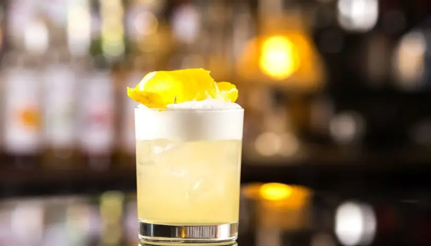 Whisky Sour z pianką i skórką cytryny w szklance na tle rozmytego baru.