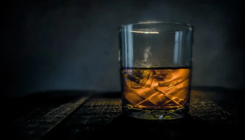 Szklanka bursztynowego trunku z lodem na drewnianym blacie. Z czego robi się whisky? Z ziaren zbóż, wody i drożdży, a potem leżakuje w beczkach.