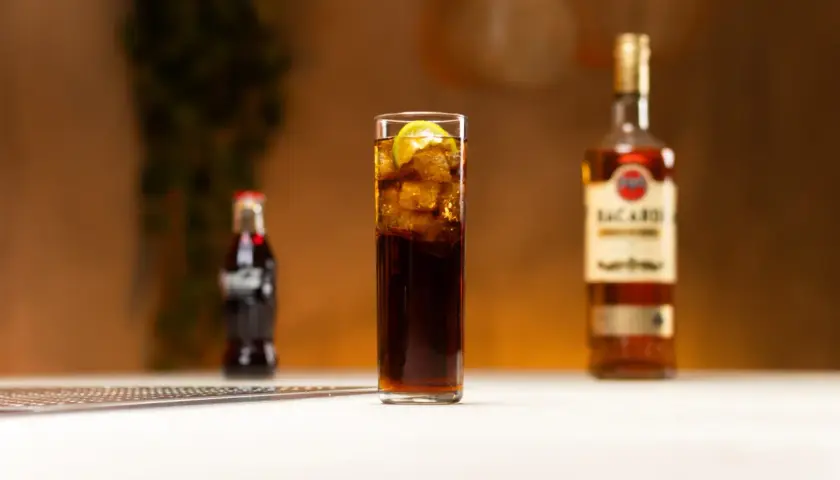 Szklanka z drinkiem z rumem Bacardi, lodem i limonką, obok butelka Coca-Coli i rumu.