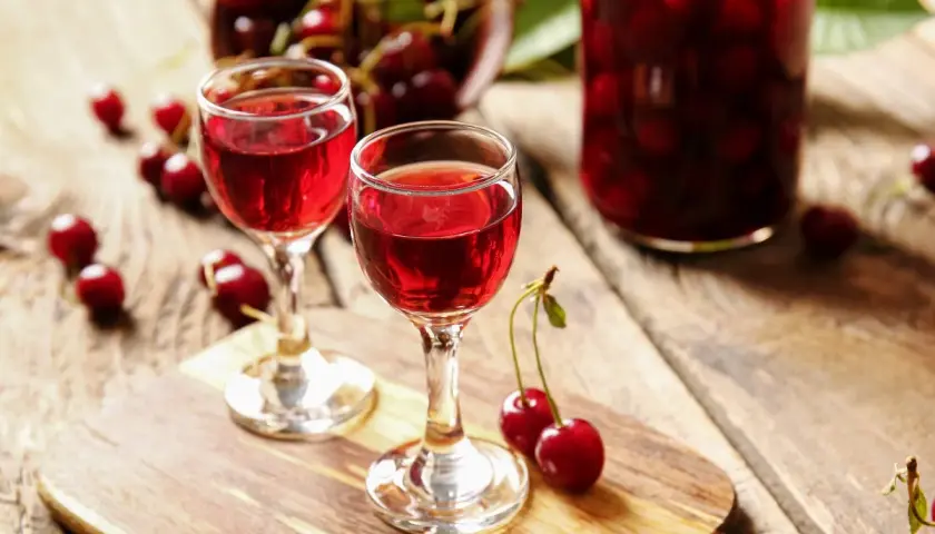 Dwa kieliszki z rubinowym winem z czereśni, obok świeże owoce i słoik pełen wiśni.
