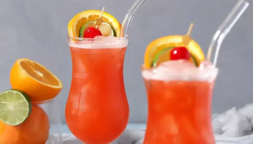 Dwa drinki z rumem, ozdobione plasterkami pomarańczy, limonki i wiśnią, z owocami w tle.