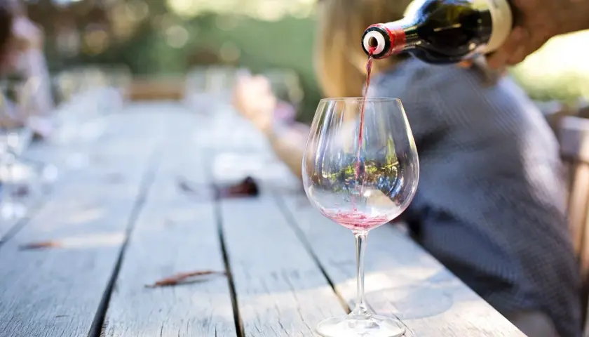 Czerwone wino Carlo Rossi nalewane do kieliszka na drewnianym stole, w tle rozmyte postacie i zieleń.